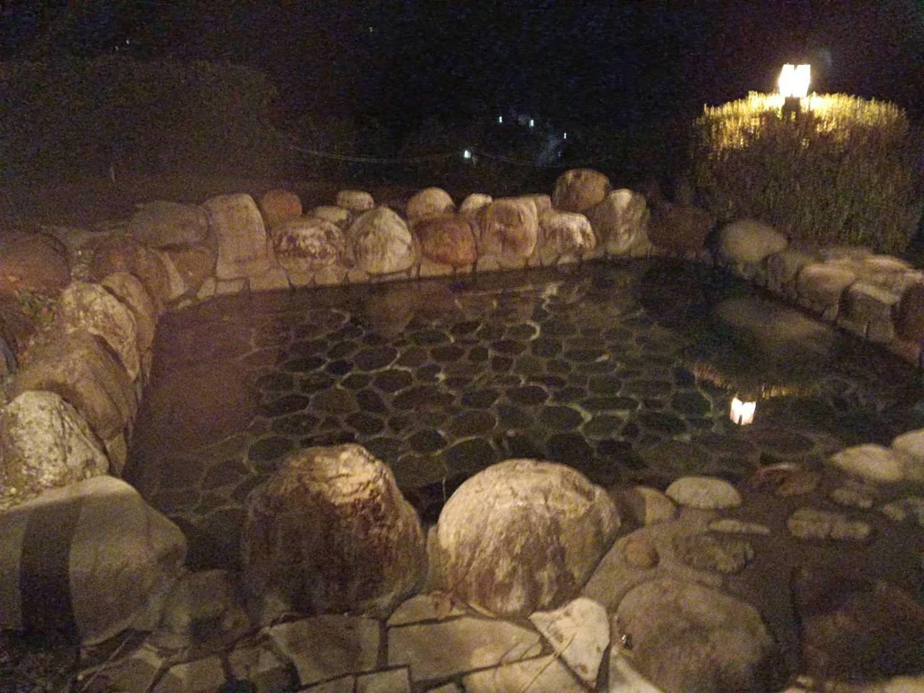 夜の露天風呂。丸い石を積んだ縁に囲まれ、生垣を照らす灯りが湯面に映る